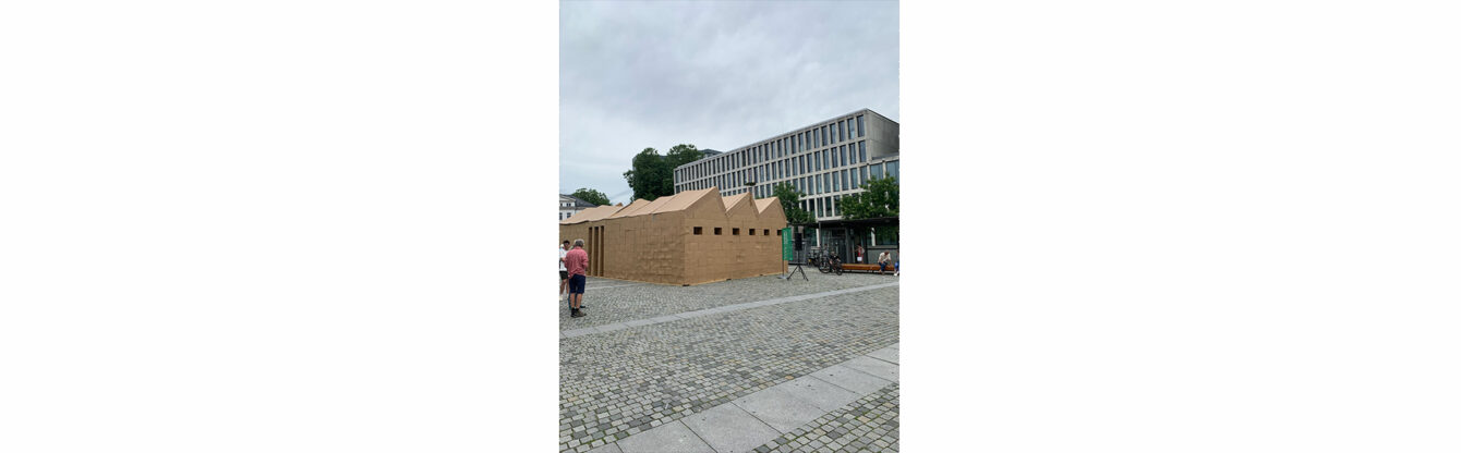 Das Bild zeigt die Kunsthalle Göppingen, nachgebaut aus braunem Karton, die auf dem gepflasterten Bahnhofsvorplatz in Göppingen steht. Das Gebäude hat mehrere kleine Fenster und ein gezacktes Dach. Im Hintergrund ist ein modernes, graues Gebäude mit mehreren Stockwerken und großen Fenstern zu sehen. Ein bewölkter Himmel vervollständigt die Szenerie, während einige Menschen in der Nähe des Kartongebäudes stehen.