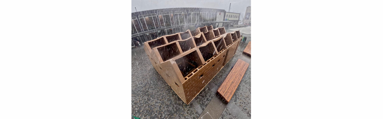 Das Bild zeigt die NEOKunsthalle Frank Bölter aus Karton, die auf dem gepflasterten Bahnhofsvorplatz steht und teilweise eingestürzt ist. Die wellenförmigen Dachelemente sind stark beschädigt und haben sich nach innen verbogen. Es scheint stark zu regnen, was wahrscheinlich zum Verfall des Kartongebäudes beigetragen hat. Einige Menschen stehen in der Nähe, während die Szene eine verregnete, stürmische Atmosphäre vermittelt.